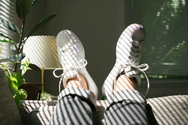 Feet wearing sneakers resting on the end of a sofa with lamp and plant behind