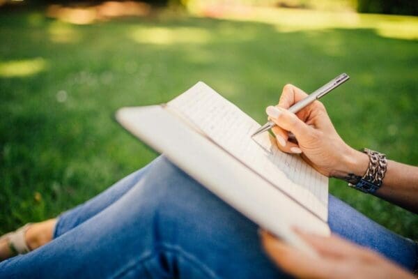 Hand of woman writing in notebook on her lap while sitting on the grass