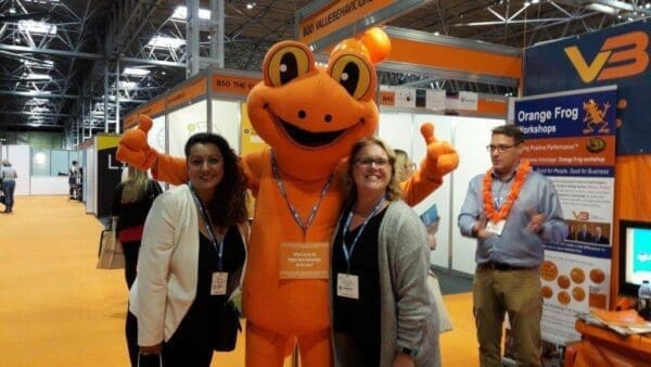 Orange frog mascot with presenters at the World of Learning Expo