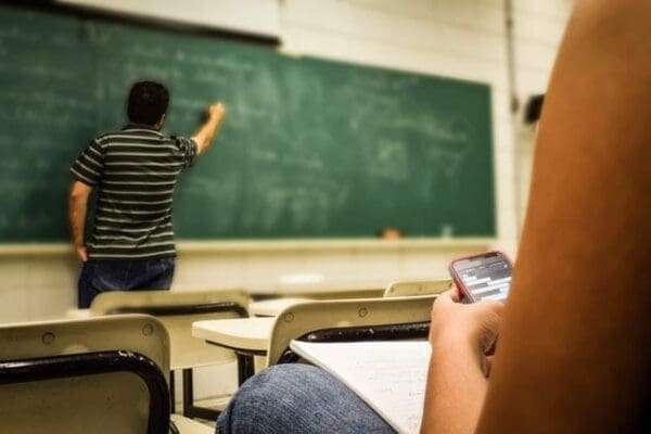 Student typing on phone whilst teacher writes on chalkboard