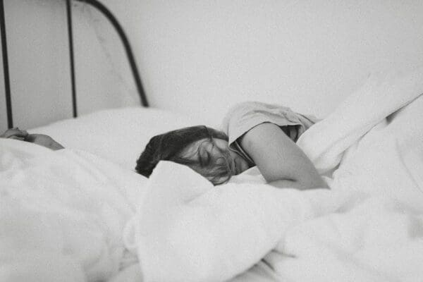 Black and white image of a woman sleeping in a white bed to increase her productivity