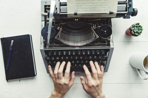 Hands typing on an old-fashioned typewriter with notepad next to it