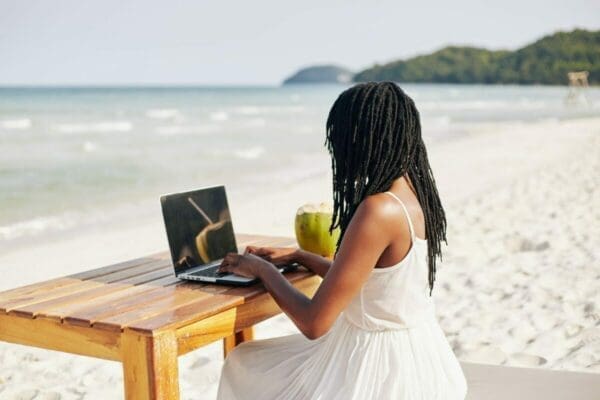 Black lady working remotely with her laptop and desk at the beach