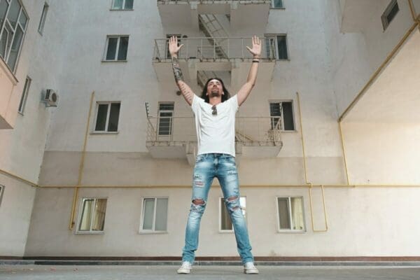 Man wearing a white shirt and jeans standing in front of a building white hands up