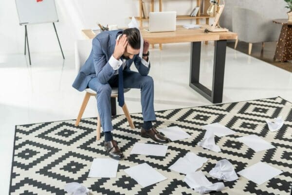 Businessman sitting on chair with hands over his ears and looking at papers on floor is facing pressure