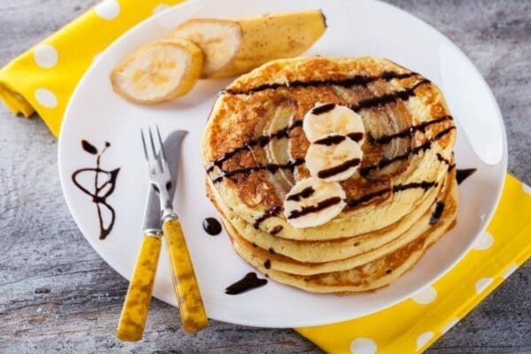Stacked banana pancake with chocolate syrup and yellow utensils