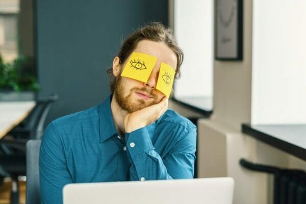 Tired sleeping young male office worker hiding eyes with funny sticky notes