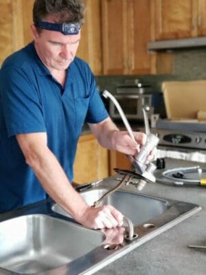 Handyman in a blue shirt with a forehead light is repairing a kitchen sink 