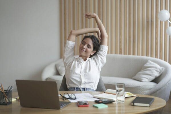 Tired italian lady remote employee stretching arms while sitting at desk with laptop
