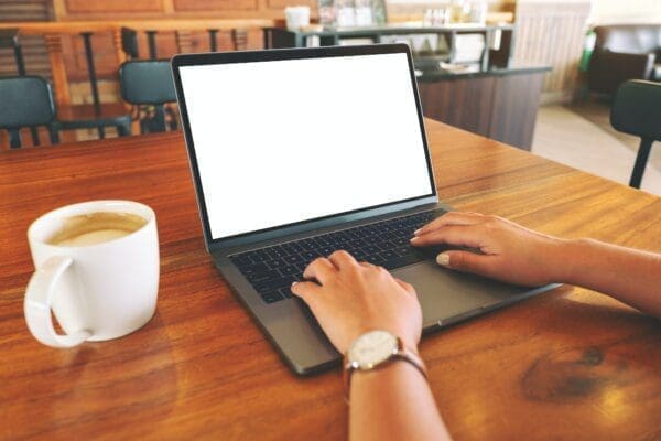 Person typing on a laptop with a blank white screen around a wooden table with a white coffee cup