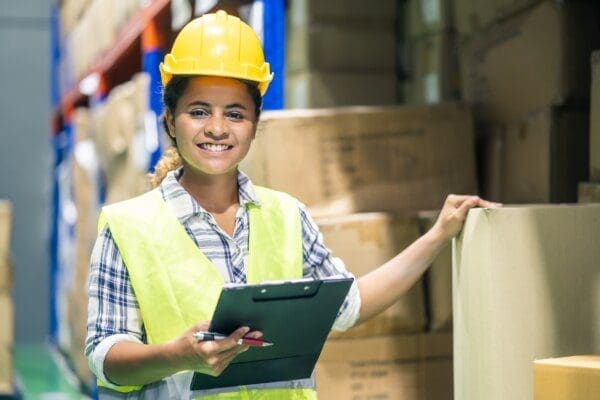 Female worker waering safety helmet and vest checking boxes in stock in a warehouse