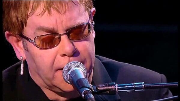 Close up of Elton John singing at the piano into a microphone while wearing brown glasses