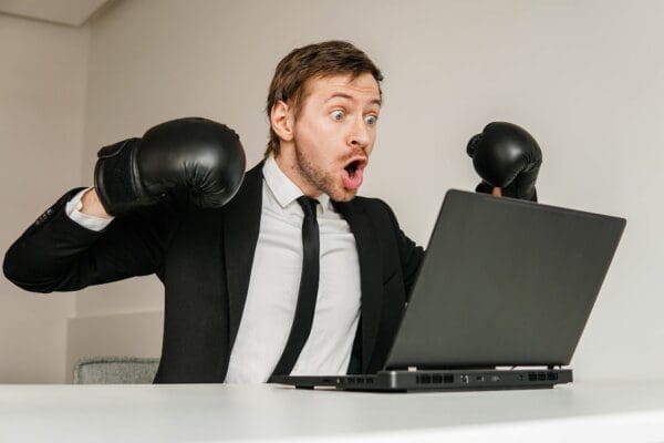 Young angry businessman in black suit wearing boxing gloves using laptop