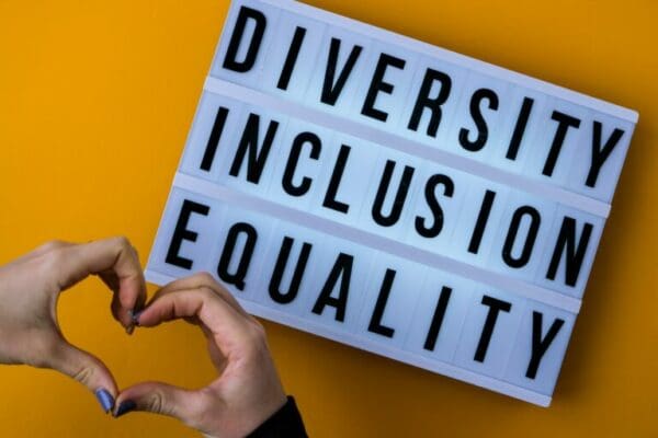 Diversity inclusion equality lettering on orange background next to a heart-shaped hand sign