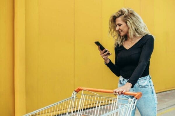 Woman using smartphone while holding shopping cart with yellow backgorund