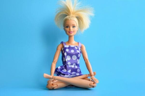 A Barbie doll in a lilac dress with white polka dots sits in a yoga pose with her legs crossed