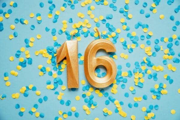 Number 16 sixteen golden celebration birthday candle on yellow and blue confetti Background